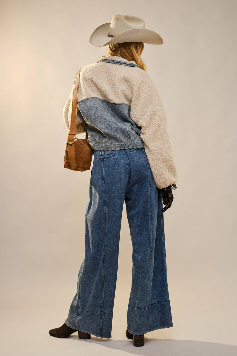 Person wearing a two-tone jacket, jeans, and cowboy hat with a brown bag on a plain background