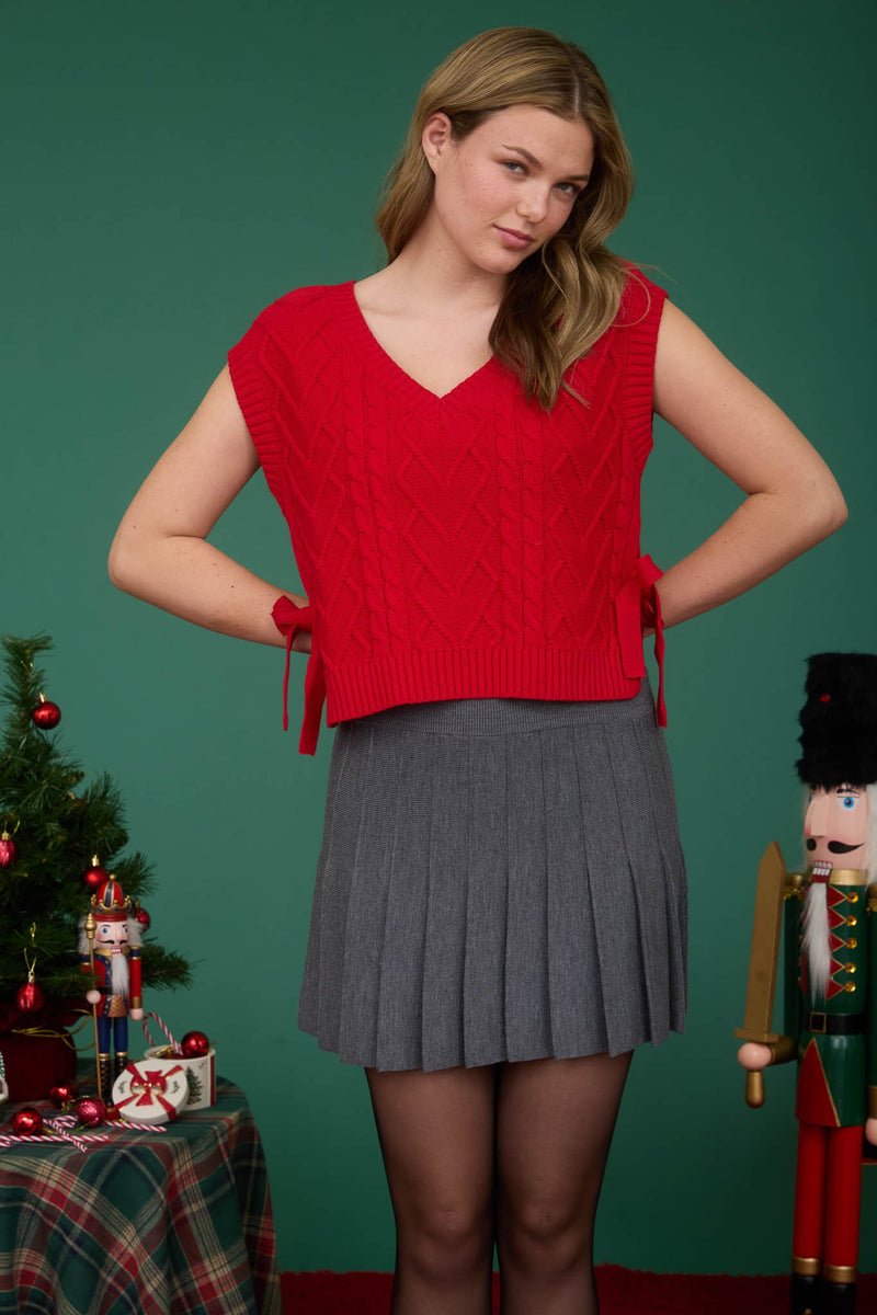 Vibrant red cable knit V-neck sweater vest with side ties, paired with a pleated gray skirt, perfect for Long Island boutique fashion.