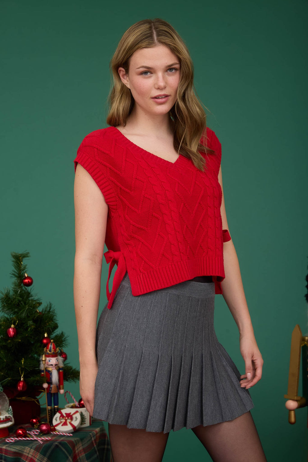 Vibrant red cable knit V-neck sweater vest with side ties, paired with a pleated gray skirt, perfect for Long Island boutique fashion.