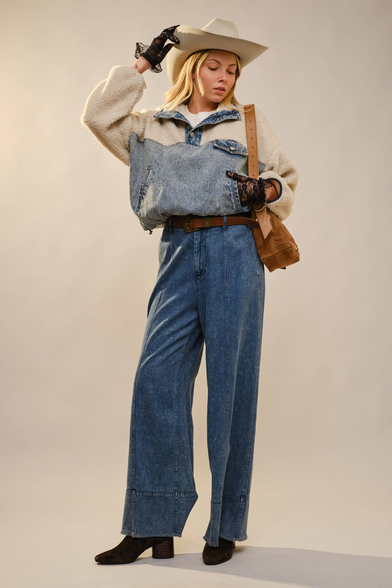 Person wearing a denim outfit with a beige sweater, wide-brimmed hat, and holding a brown bag on a plain background.